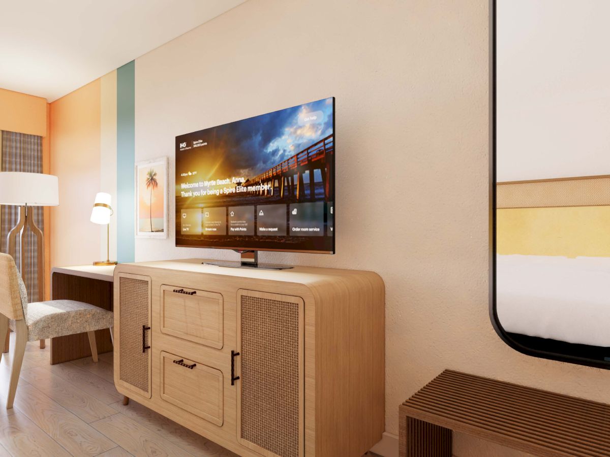 The image shows a modern hotel room with a TV on a wooden stand, a desk, chair, lamp, and a partial view of the bed and balcony.