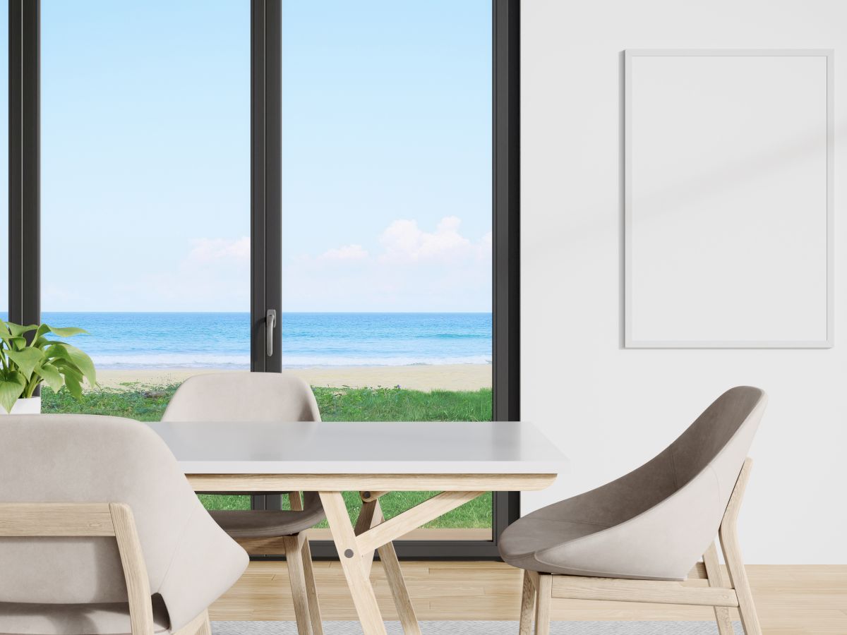 A modern room with a table and chairs, a plant, and a large window showing a beach view. There's also a blank white frame on the wall.